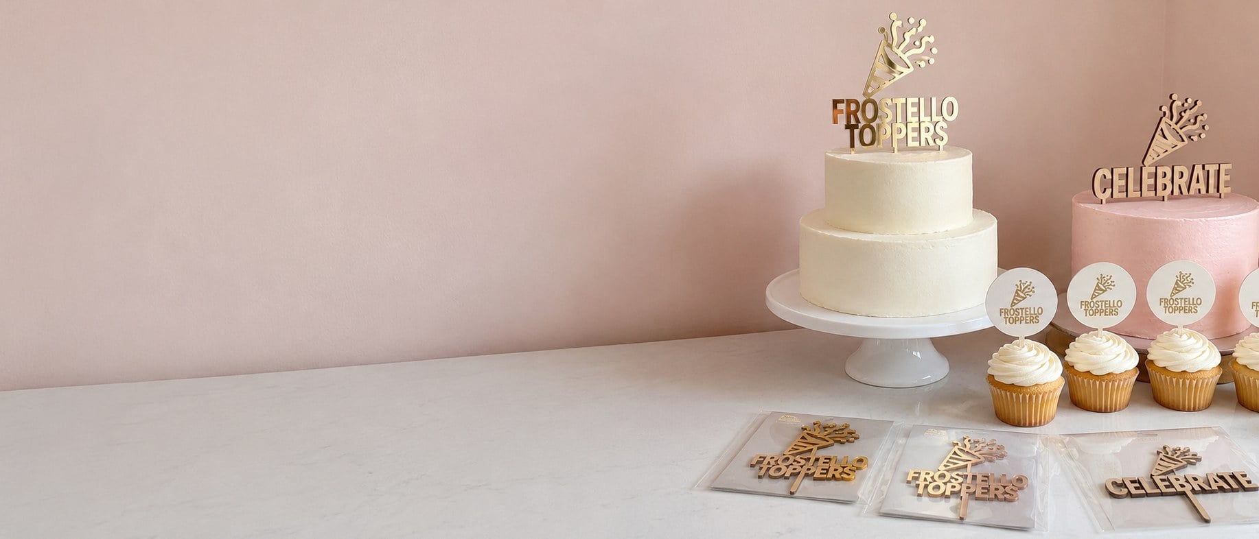 Custom acrylic, wooden and mirror gold-tone cake toppers on a white cake and cupcakes against a plain wall